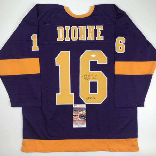 Flat lay of signed purple and gold LA Kings hockey jersey with number 16, "DIONNE" nameplate, and JSA authentication certificate attached.