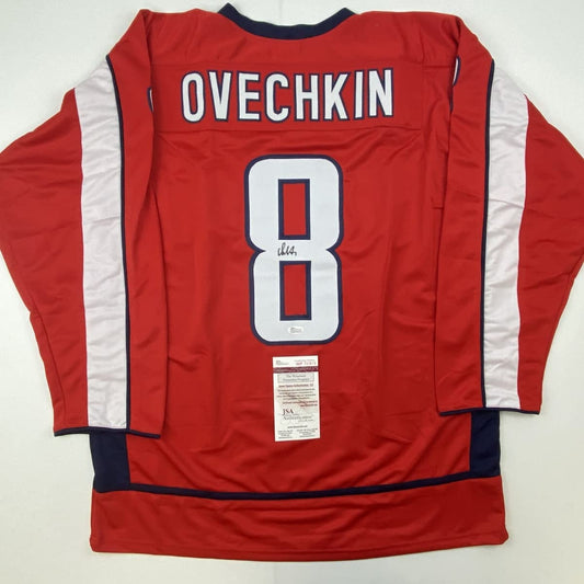 Back view of red hockey jersey with white number 8, "OVECHKIN" nameplate, white side stripes, and JSA authentication certificate attached.