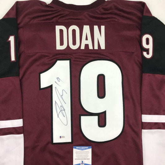Back view of maroon and black hockey jersey with white number 19, "DOAN" nameplate, and autographed signature on the number, displaying Beckett authentication certificate.