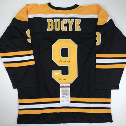 Back view of black Boston Bruins hockey jersey number 9 with yellow and white stripes, signed by Johnny Bucyk with HOF 1981 notation and JSA authentication label.