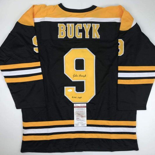 Back view of black Boston Bruins hockey jersey number 9 with yellow and white stripes, signed by Johnny Bucyk with HOF 1981 notation and JSA authentication label.
