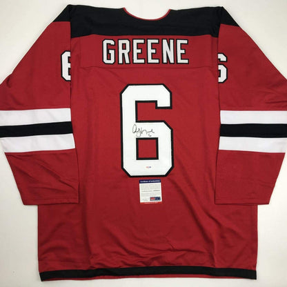 Red hockey jersey with black and white stripes displaying "GREENE" and number 6, signed, flat lay view with authenticity certificate visible.