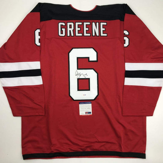 Red hockey jersey with black and white stripes displaying "GREENE" and number 6, signed, flat lay view with authenticity certificate visible.