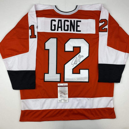 Flat lay of red, white, and black striped hockey jersey number 12 with "GAGNE" nameplate and signature, JSA authentication label visible.