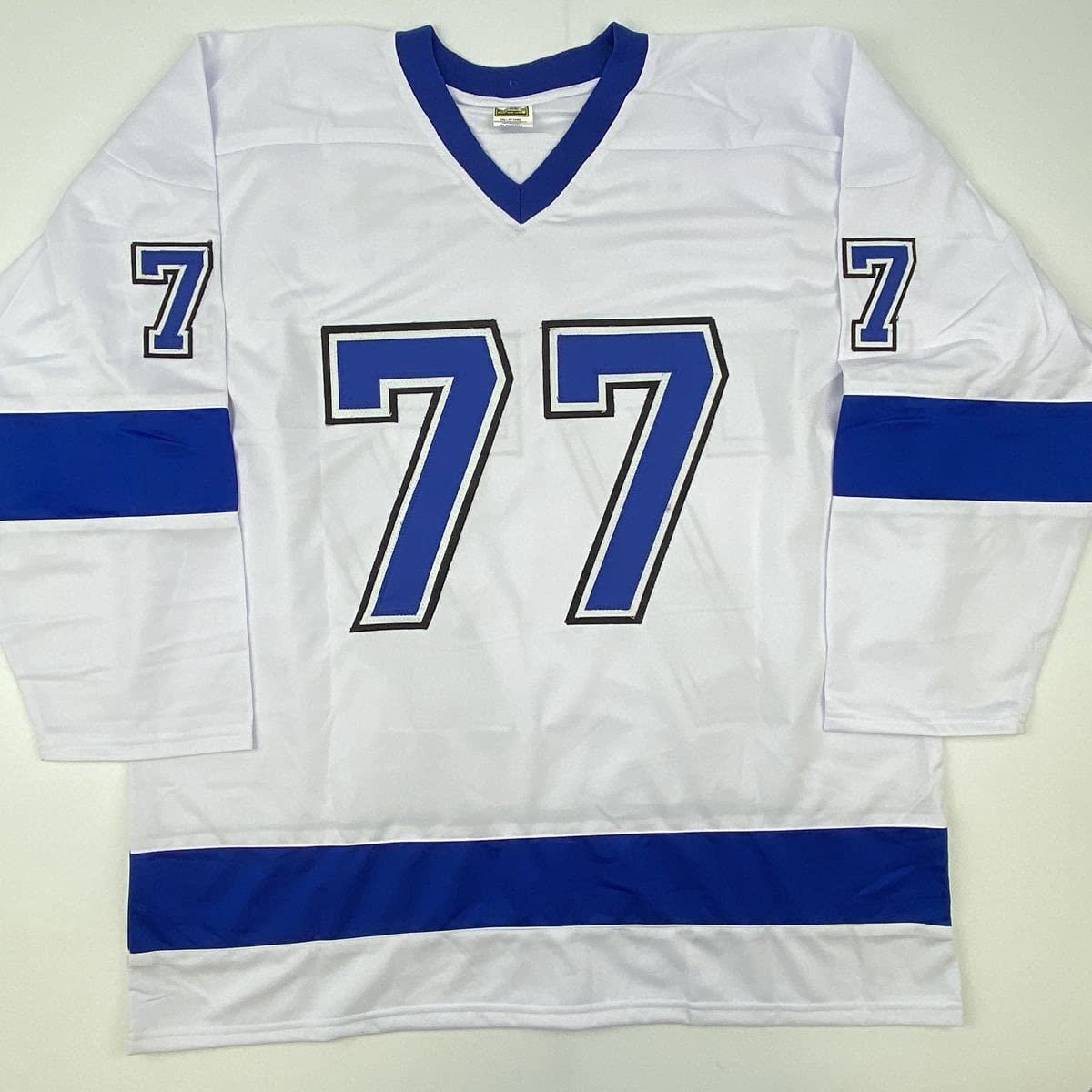 White hockey jersey with blue v-neck collar, blue horizontal stripes, and large blue number 77 with black outline displayed flat on light background.