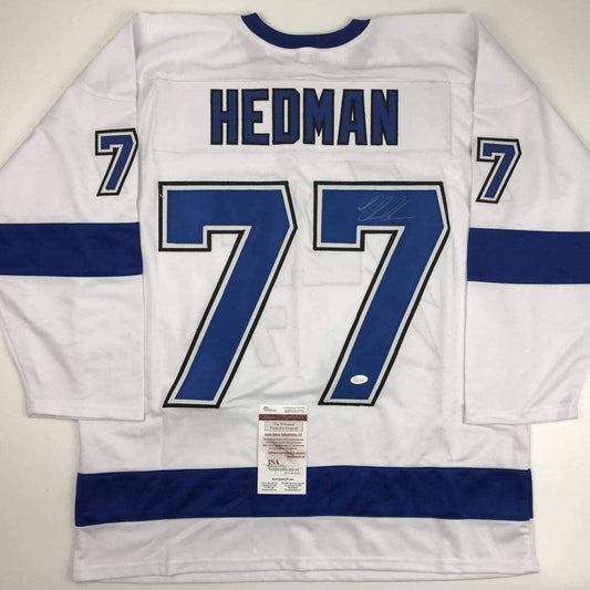 White hockey jersey with blue sleeves and stripes displaying "HEDMAN" and number 77, with JSA authentication certificate visible at center bottom.