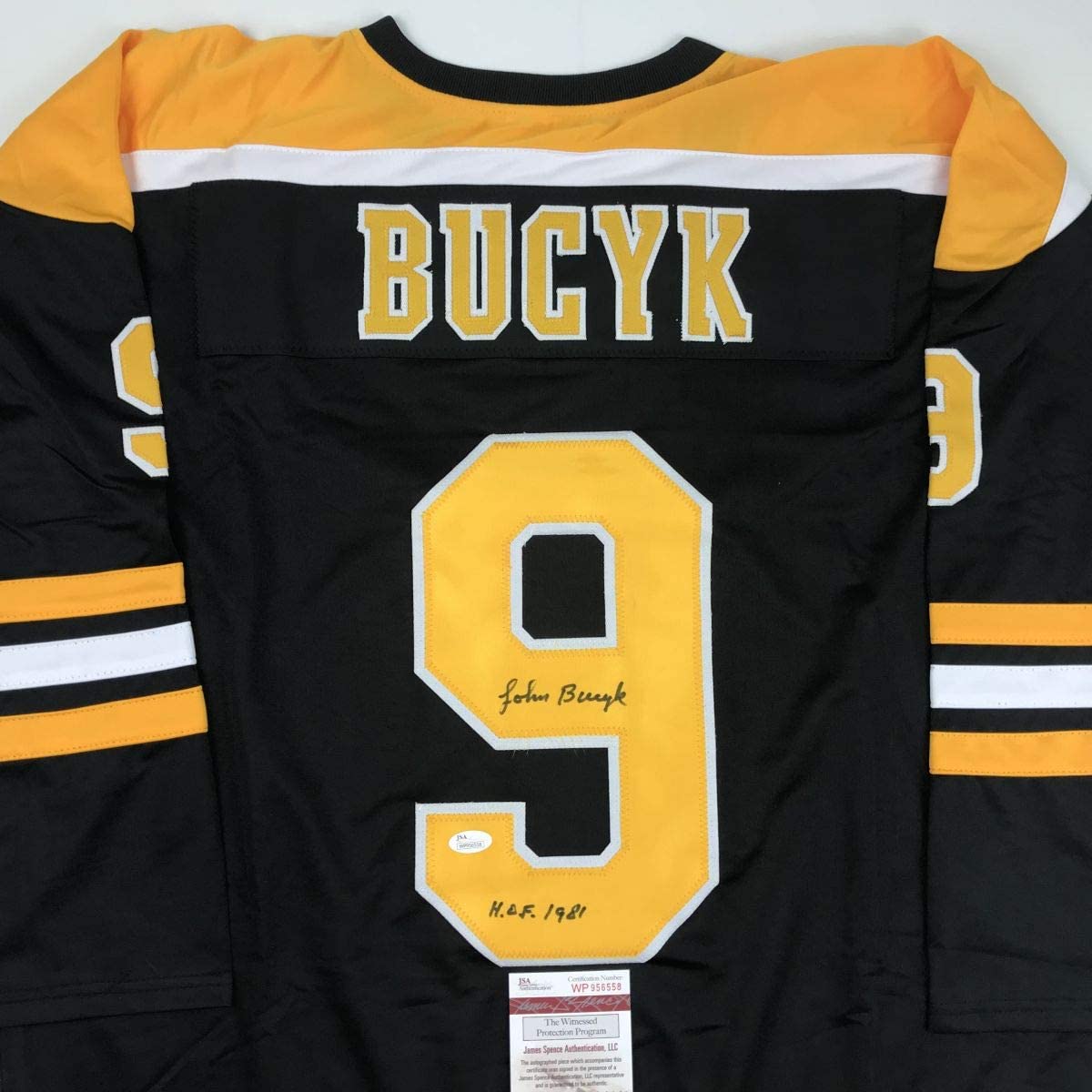Back view of black Boston Bruins hockey jersey number 9 with yellow and white trim, signed by Johnny Bucyk with HOF 1981 notation and JSA authentication label.