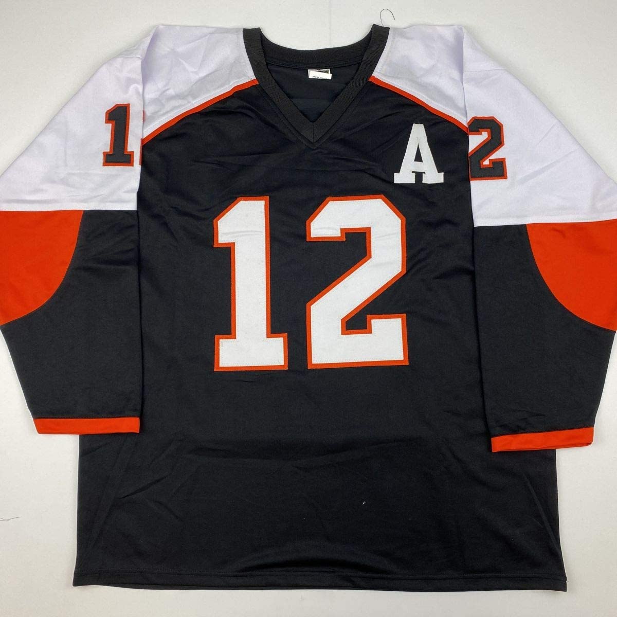 Black hockey jersey with number 12, white and orange trim, displayed flat on hanger against white background.