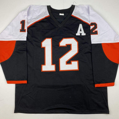 Black hockey jersey with number 12, white and orange trim, displayed flat on hanger against white background.