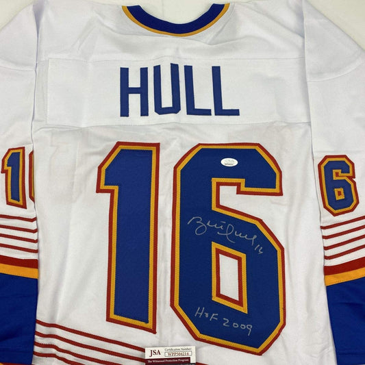 Close-up of white hockey jersey front showing "HULL" nameplate, blue number 15 with signature and "HOF 2009" inscription, and JSA authenticity label.