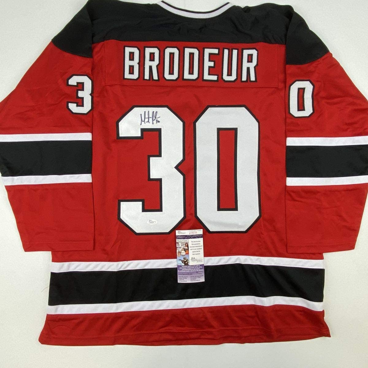 Red New Jersey Devils hockey jersey with "BRODEUR" and number 30 displayed on back, featuring black and white stripes, with JSA authentication certificate visible.