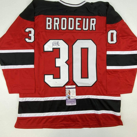 Red New Jersey Devils hockey jersey with "BRODEUR" and number 30 displayed on back, featuring black and white stripes, with JSA authentication certificate visible.