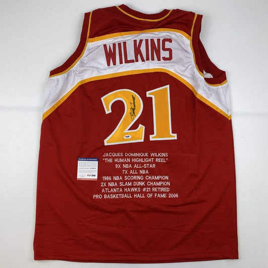 Back view of red Atlanta Hawks jersey #21 with "WILKINS" nameplate, gold and white trim, and white stat text detailing Jacques Dominique Wilkins' career achievements.