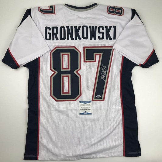 Back view of white New England Patriots football jersey number 87 with "GRONKOWSKI" nameplate, navy sleeves, red trim, and Beckett authentication certificate visible.