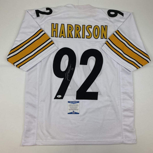 White Pittsburgh Steelers football jersey number 92 with "HARRISON" nameplate, gold and black striped sleeves, signed with Beckett authentication sticker, flat lay back view.