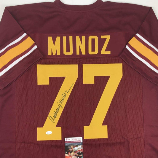Back view of maroon USC football jersey #77 with "MUNOZ" nameplate and handwritten signature, displaying JSA authenticity certificate label.