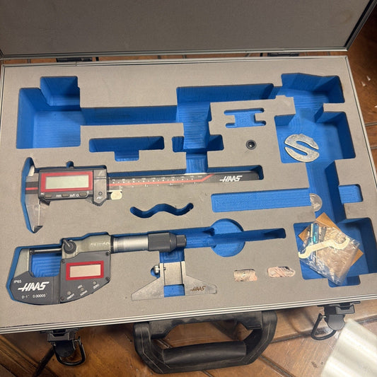 Open aluminum case displaying blue foam-lined compartments holding two digital micrometers with red display screens, rulers, and loose metal parts.