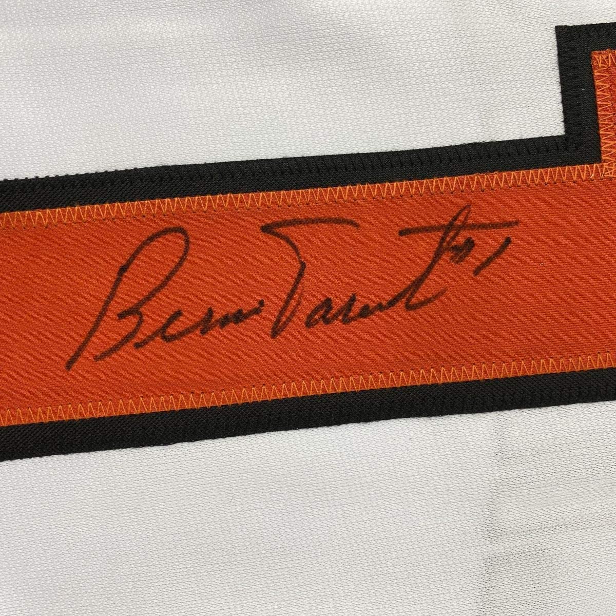 Close-up of handwritten signature "Bernie Parent" in black ink on orange and black striped ribbon trim against white fabric background.