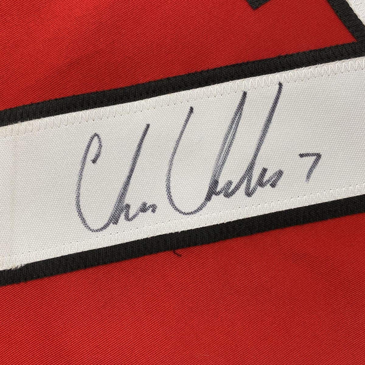 Close-up of signature on white stripe of red and black hockey jersey reading "Chris Chelios 7".