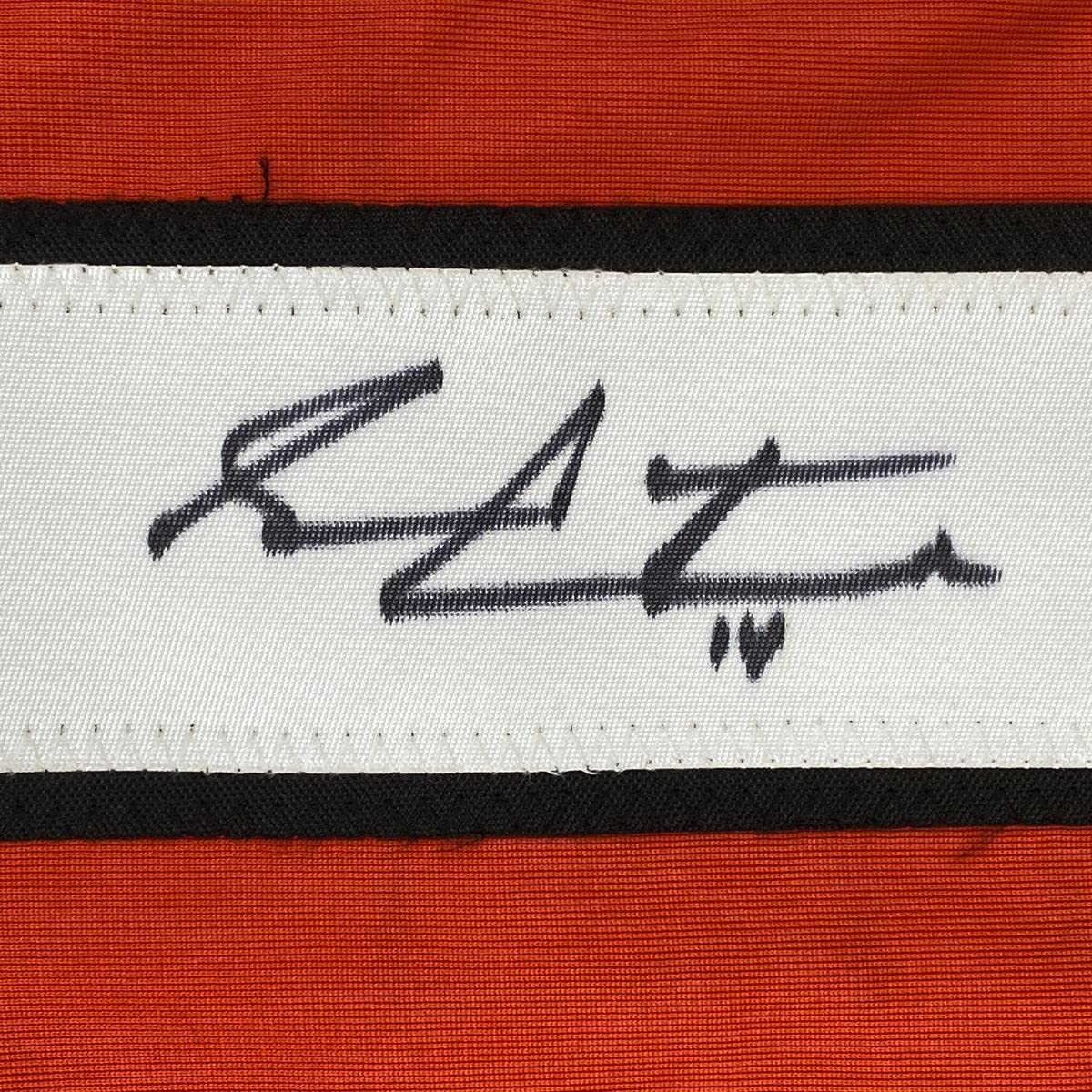 Close-up of a handwritten signature in black ink on a white fabric stripe between black and orange horizontal bands.
