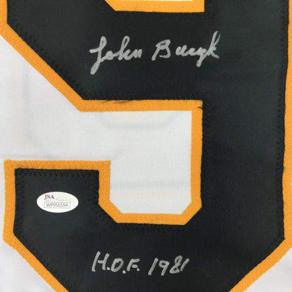 Close-up of black hockey jersey back with gold trim, white pocket, handwritten signature "John Bucyk" and "H.O.F 1981" text, JSA authentication sticker visible.