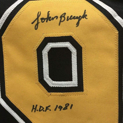 Close-up of signed yellow hockey jersey patch showing number 0 with white and black trim, signed "John Bucyk" at top and "H.D.F. 1981" at bottom.
