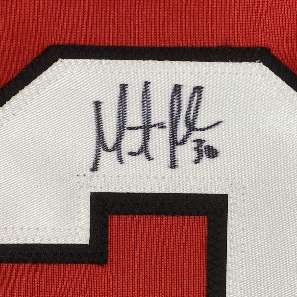 Close-up of handwritten signature "Marty Brodeur" in black ink on white jersey label against red fabric background.