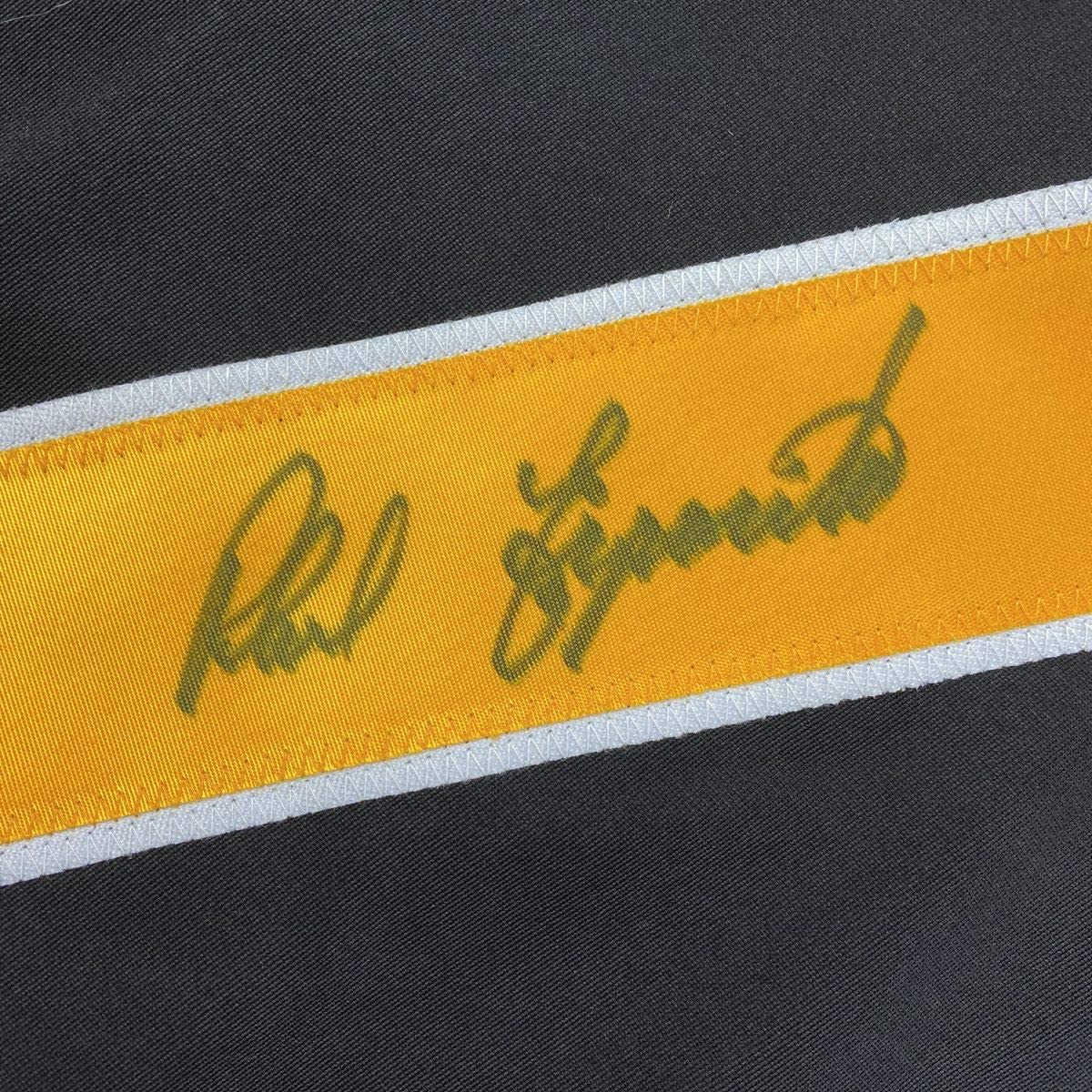Close-up of autographed yellow and white stripe on black Boston Bruins jersey with green ink signature.