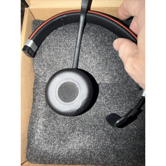 Overhead view of black Jabra Evolve 65 SE headset with red accent stripe lying on gray fabric packaging inside cardboard box.
