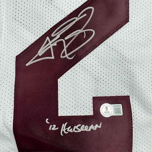 Close-up of maroon jersey sleeve showing autographed signature and "12 Heisman" text with authentication hologram sticker on white mesh fabric.