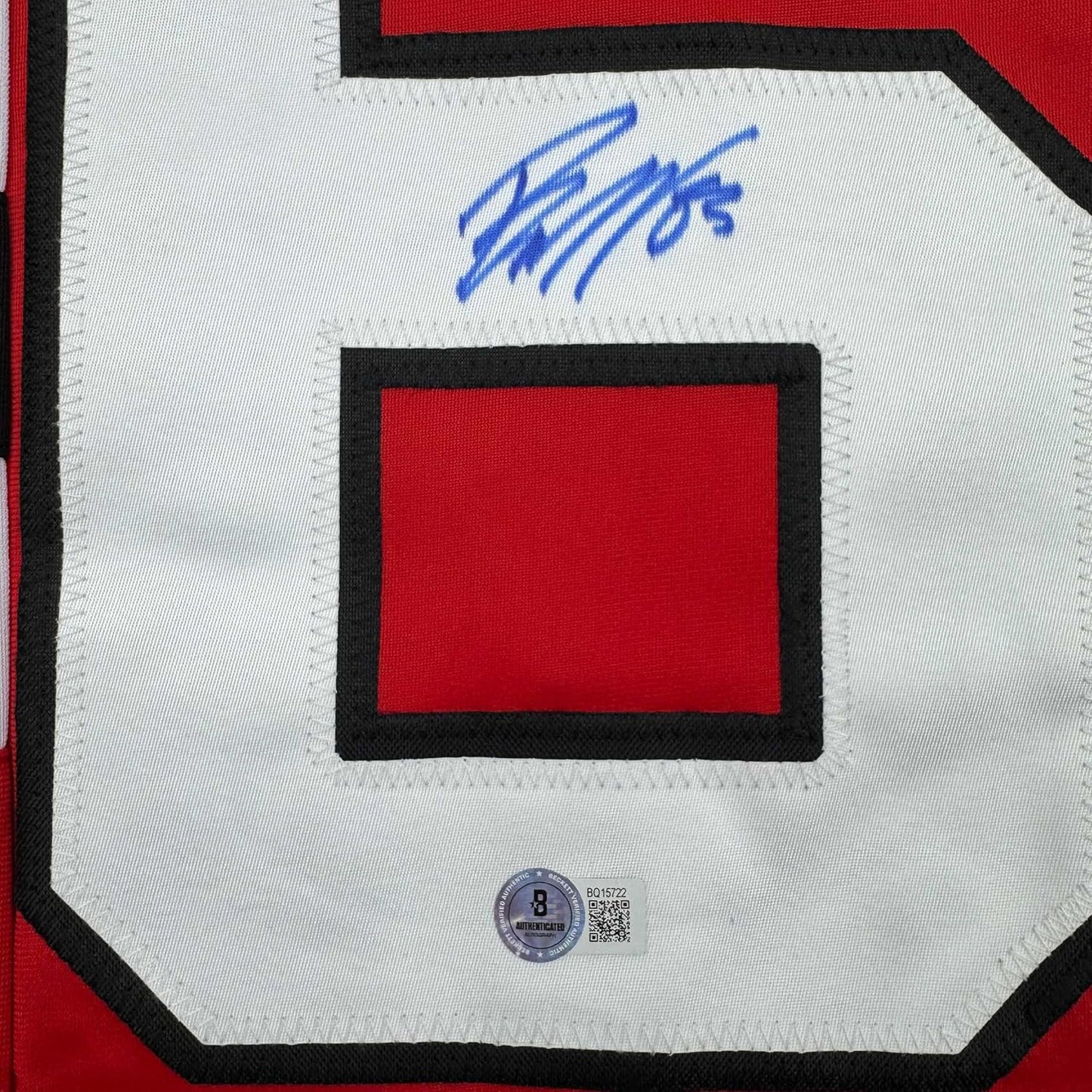 Close-up of blue autograph signature on white jersey with red rectangular patch and black border, Beckett authentication hologram visible at bottom.