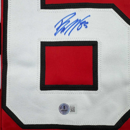 Close-up of blue autograph signature on white jersey with red rectangular patch and black border, Beckett authentication hologram visible at bottom.