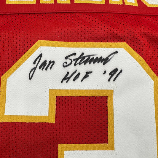 Close-up of signed nameplate on red jersey showing "Jan Stenerud HOF '91" in black handwriting on white label with gold border.