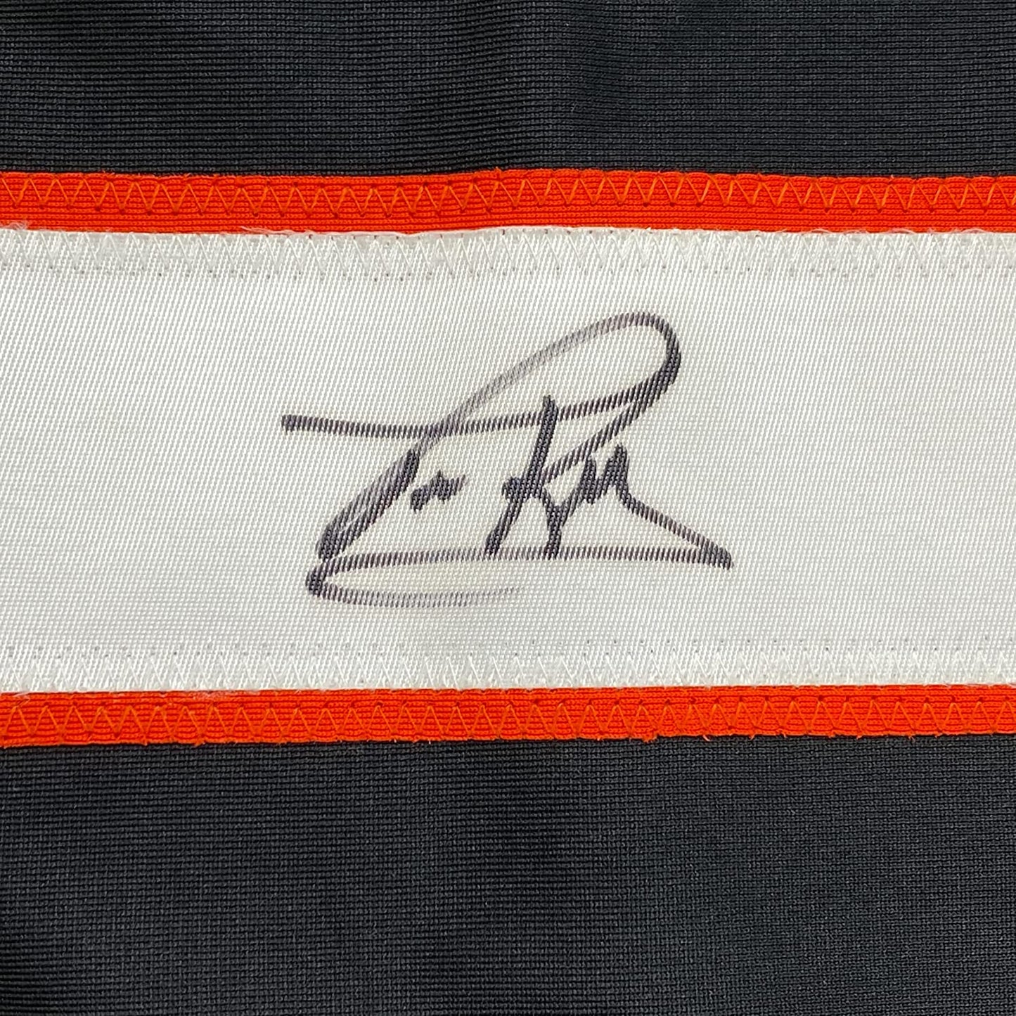 Close-up of embroidered signature on white jersey stripe between orange and black horizontal bands.