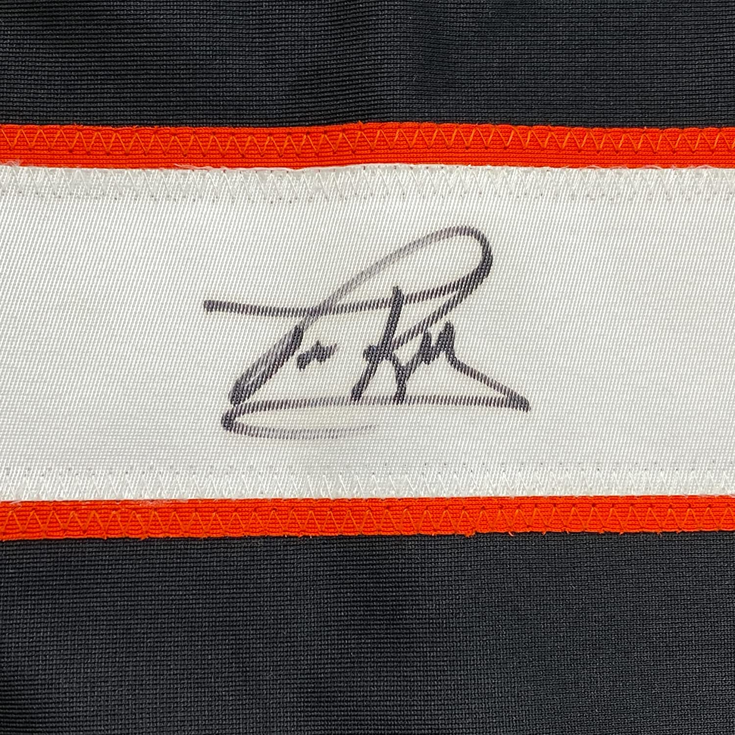Close-up of embroidered signature on white jersey stripe between orange and black horizontal bands.