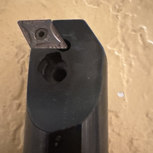 Close-up angled view of black metal boring bar tool holder showing inserted carbide insert and mounting hole against tan concrete background.