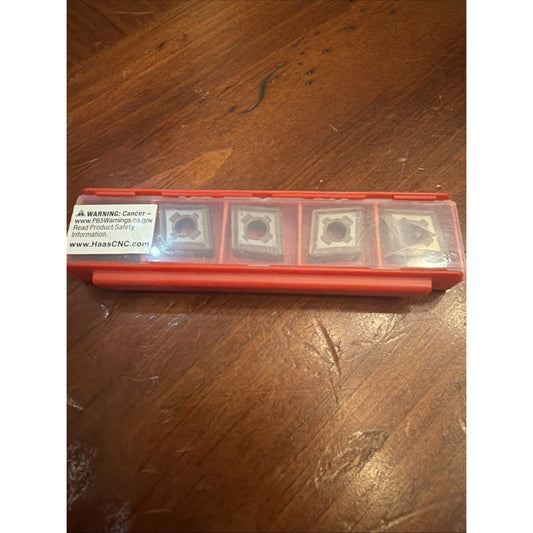 Red plastic case containing four square carbide inserts with warning label visible on wooden floor surface.