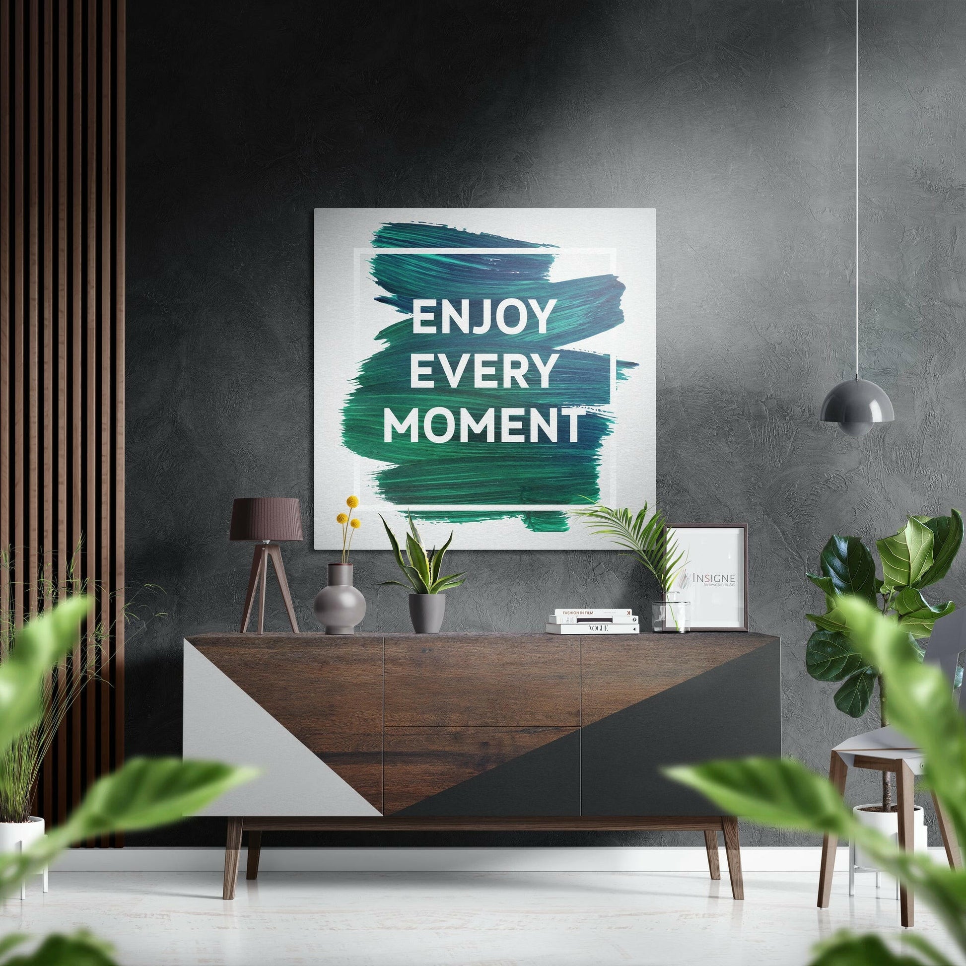 Modern living room with teal brushstroked "Enjoy Every Moment" wall art above wooden dresser with plants and pendant lamp.