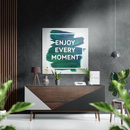 Modern living room with teal brushstroked "Enjoy Every Moment" wall art above wooden dresser with plants and pendant lamp.