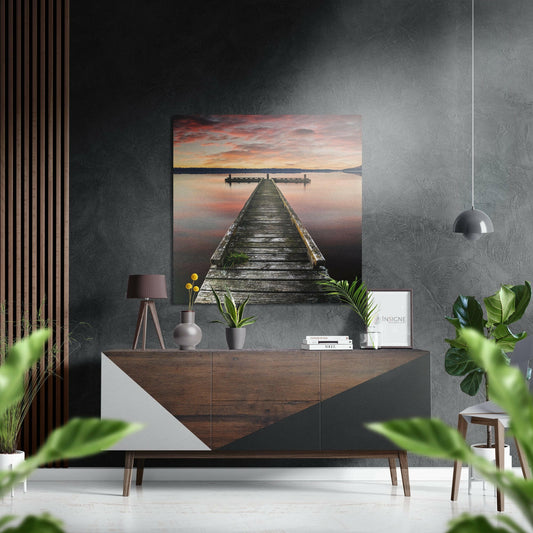 Wooden pier extending into calm water at sunset, displayed as metal wall art above a mid-century modern dresser in a styled interior.