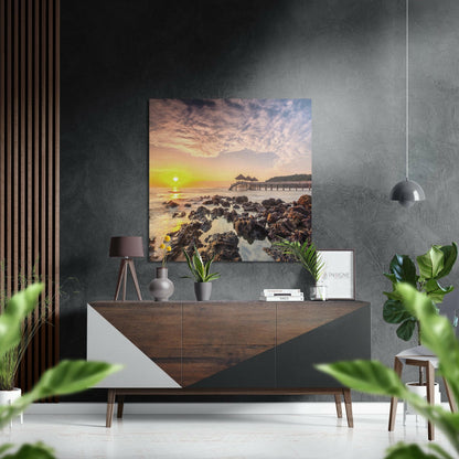 Brushed aluminum Dibond wall art displaying a golden sunset over rocky coastline with pier, mounted on dark textured wall above wooden dresser.