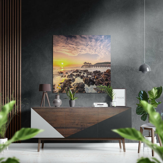 Brushed aluminum Dibond wall art displaying a golden sunset over rocky coastline with pier, mounted on dark textured wall above wooden dresser.