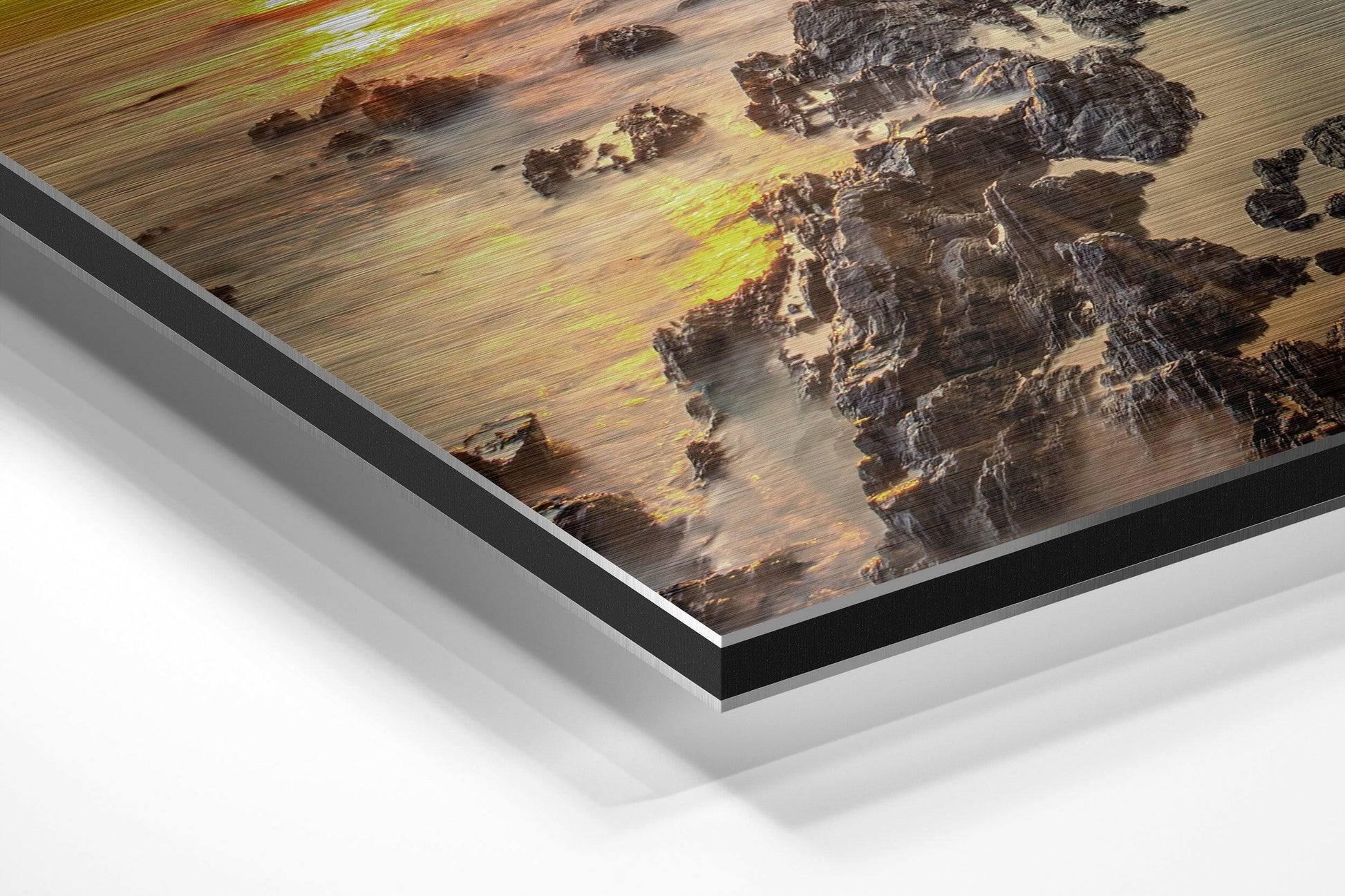 Angled perspective view of brushed aluminum Dibond print showing coastal rocks and golden sunset reflections with black and silver layered frame edges.