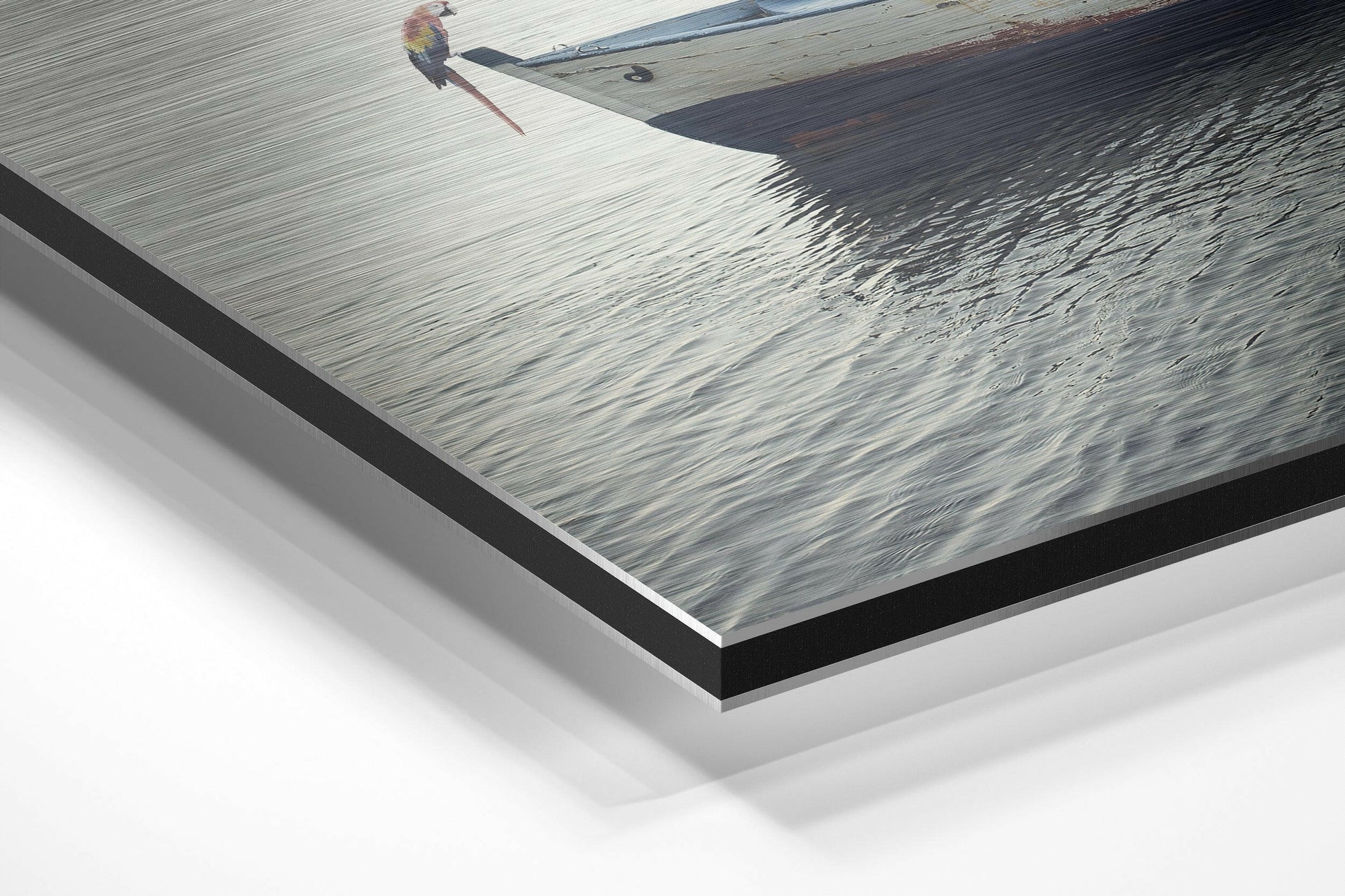 Angled view of brushed aluminum Dibond print showing a parrot perched on a wooden boat dock with calm water, with layered mounting and shadow visible.