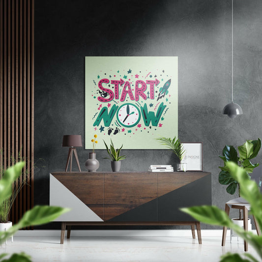 Modern interior display showing "Start Now" motivational wall art with pink text, clock, rocket, and stars on cream background, mounted above wooden console table.