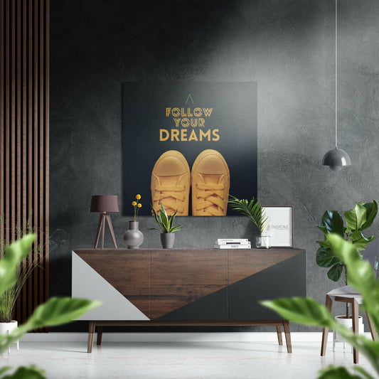 Modern interior scene featuring "Follow Your Dreams" gold text and yellow sneakers artwork on dark wall above wooden dresser with plants and pendant lamp.