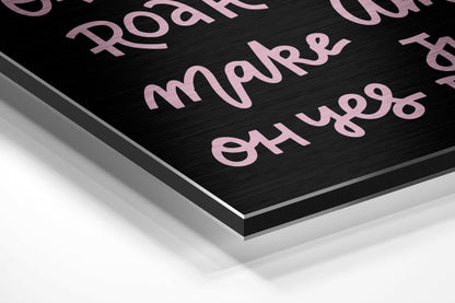 Close-up angled view of brushed black aluminum dibond panel with pink metallic text and layered edge detail against white background.