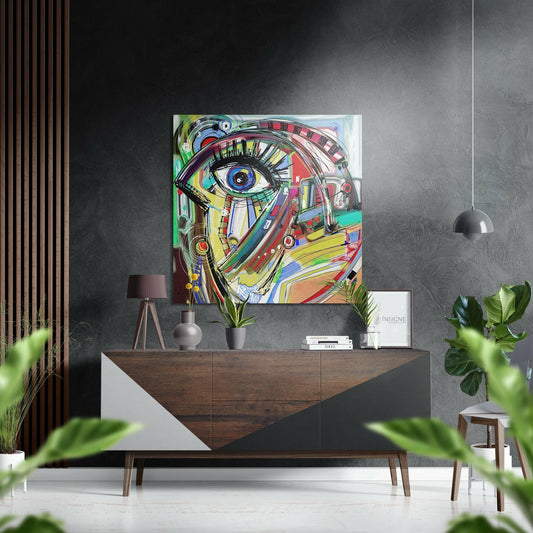 Colorful abstract blue eye art displayed on gray wall above wooden dresser with plants and pendant lamp in modern interior setting.