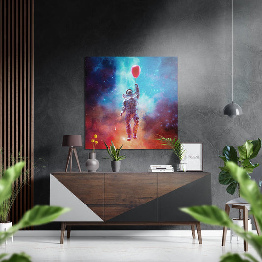 Vibrant square metal wall art depicting a spacesuited astronaut holding a red balloon against a colorful nebula backdrop, displayed above a wooden console table in a modern interior.