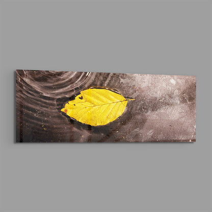Bright yellow autumn leaf floating on dark water surface, displayed as a horizontal rectangular glass wall art piece.